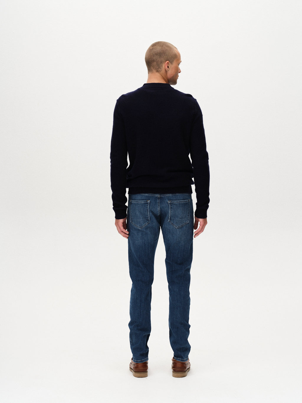 A man stands facing away from the camera wearing a dark long-sleeve shirt, brown shoes, and Gabba Jones Vanew - Mid Blue Denim jeans with a slim silhouette and super-stretch fabric for all-day comfort, set against a plain white background.