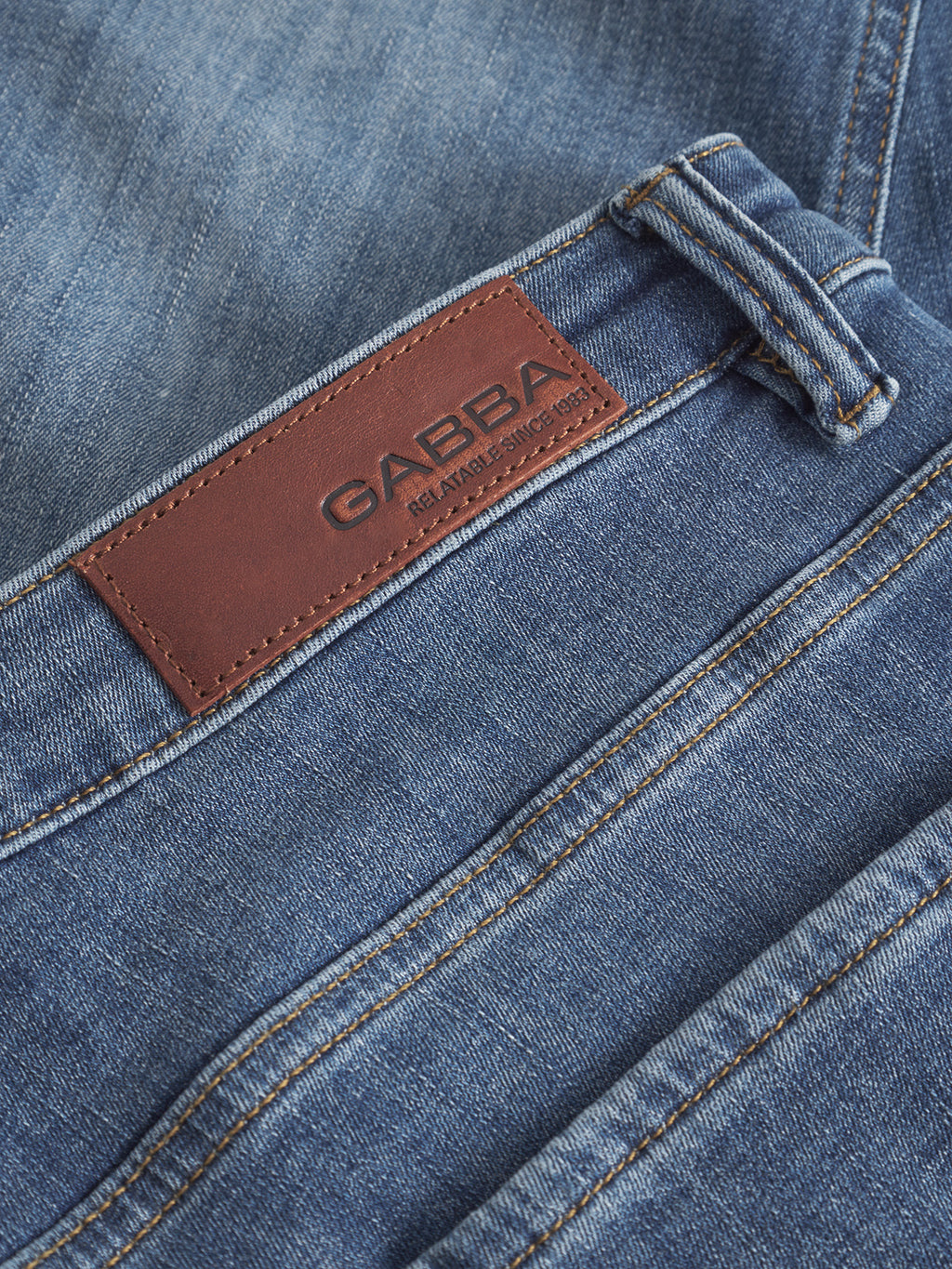 Close-up of Jones Vanew mid blue denim jeans by Gabba, showcasing a brown leather patch with GABBA and ESTABLISHED IN 1983 text. These slim silhouette jeans feature visible stitching around the belt loops and pockets, plus reinforced pockets for added durability.