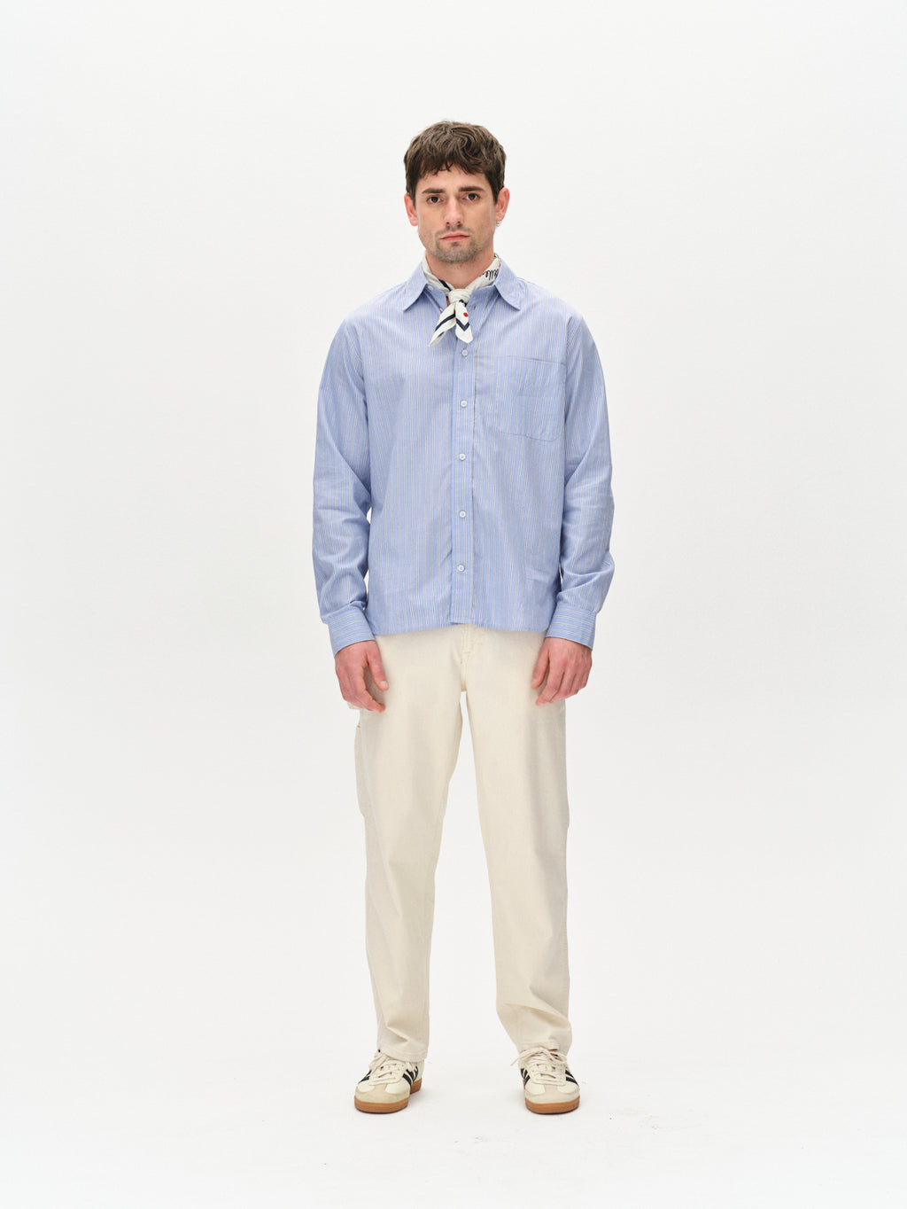 A man faces forward against a white background, wearing the Gabba Max Luis Work2 - Ecru pants, paired with a light blue slim fit shirt, white sneakers, and a multicolored scarf for modern elegance.