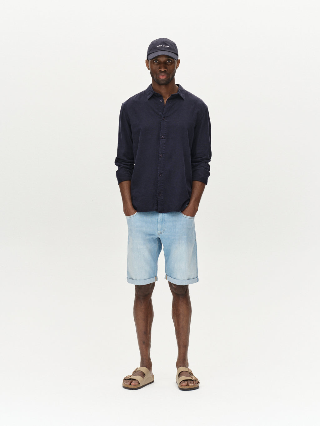A man in a dark button-up shirt confidently models the comfortable fit of Markus Selvedge Shorts Limited by Gabba in Lt. Blue Denim against a white background, accessorizing with a dark cap and brown sandals, hands casually in pockets for relaxed style.