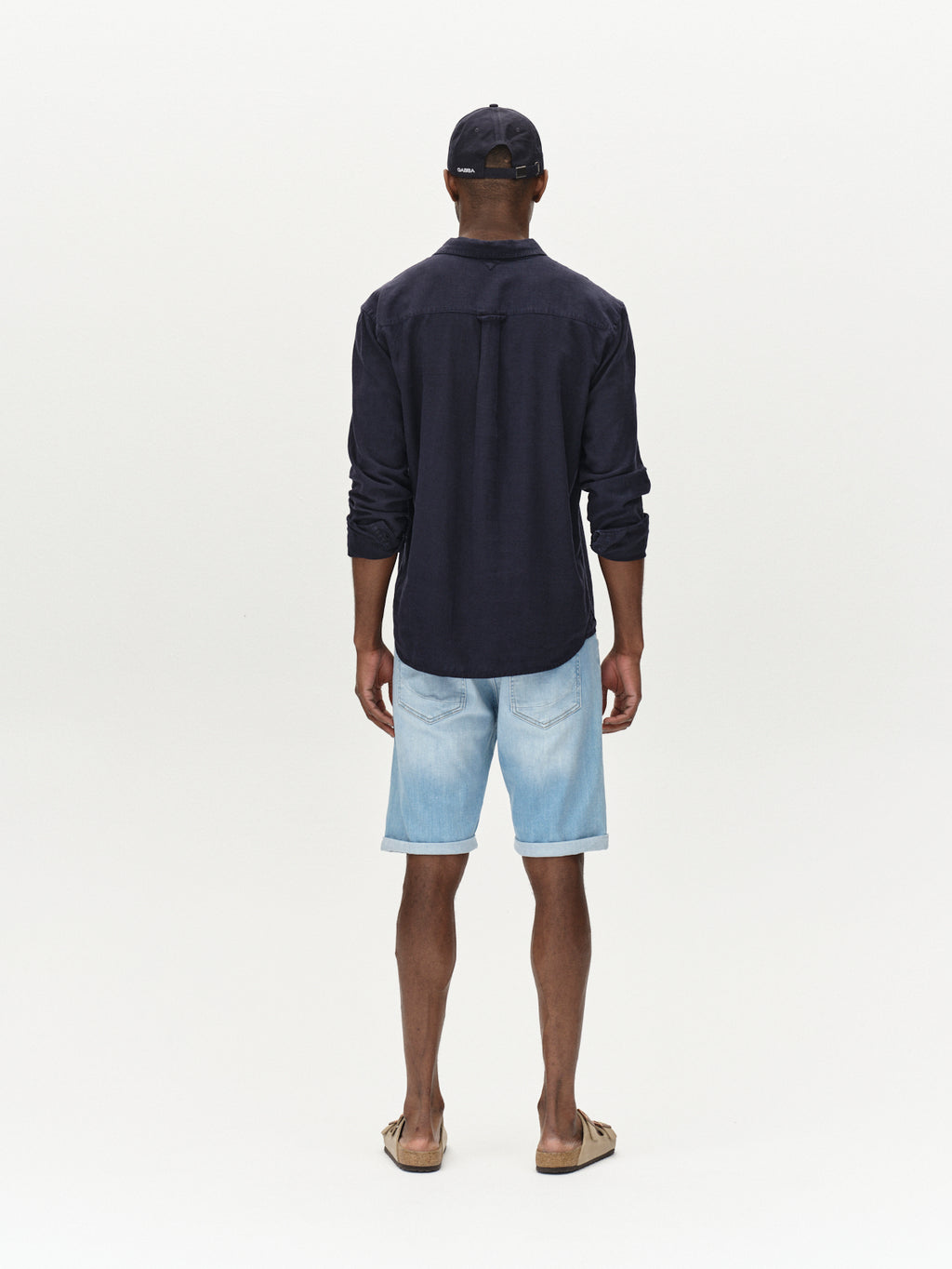 Facing away, a person wears a black baseball cap and dark long-sleeve shirt paired with Gabbas Markus Selvedge Shorts Limited in light blue denim, offering stitching details for comfort. Flip-flops complete the look against a plain white background.