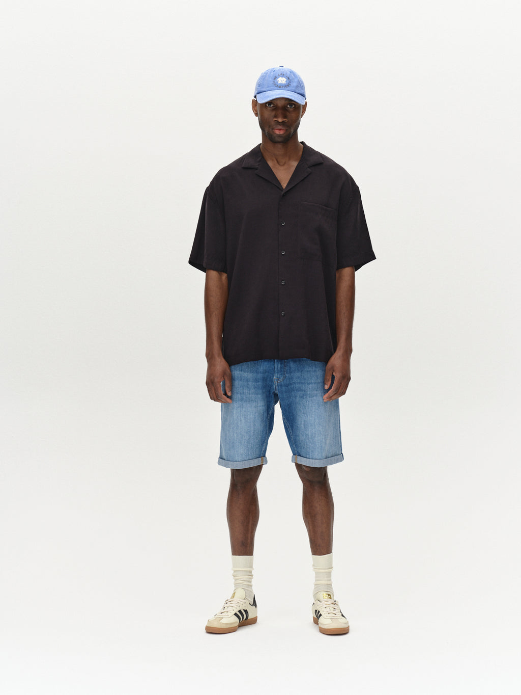 A person stands against a plain background wearing a black short-sleeve button-up shirt, Gabbas Markus Selvedge Shorts Limited in mid-blue denim, a blue baseball cap, and beige sneakers with socks.