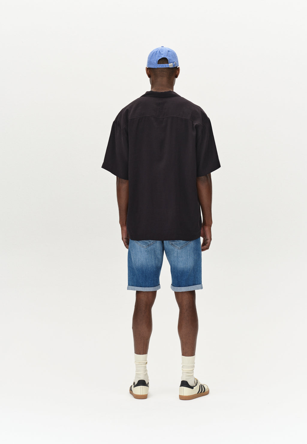 A person stands with their back to the camera, showcasing a comfortable fit in a black short-sleeve shirt and Gabbas Markus Selvedge Shorts Limited in Mid Blue Denim. Theyre wearing white socks, striped sneakers, and a light blue cap against a plain white background.