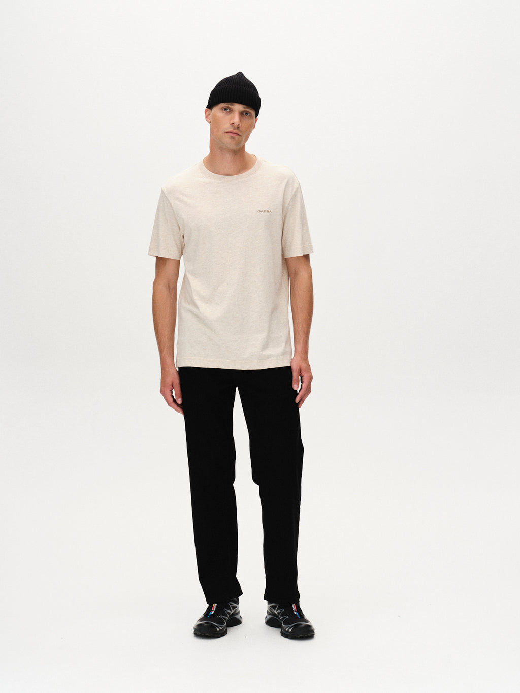 A man wearing a black beanie, black pants, and black shoes models the Gabba Dune Melange SS - Lt. Sand t-shirt against a plain white background, looking directly at the camera with a neutral expression.