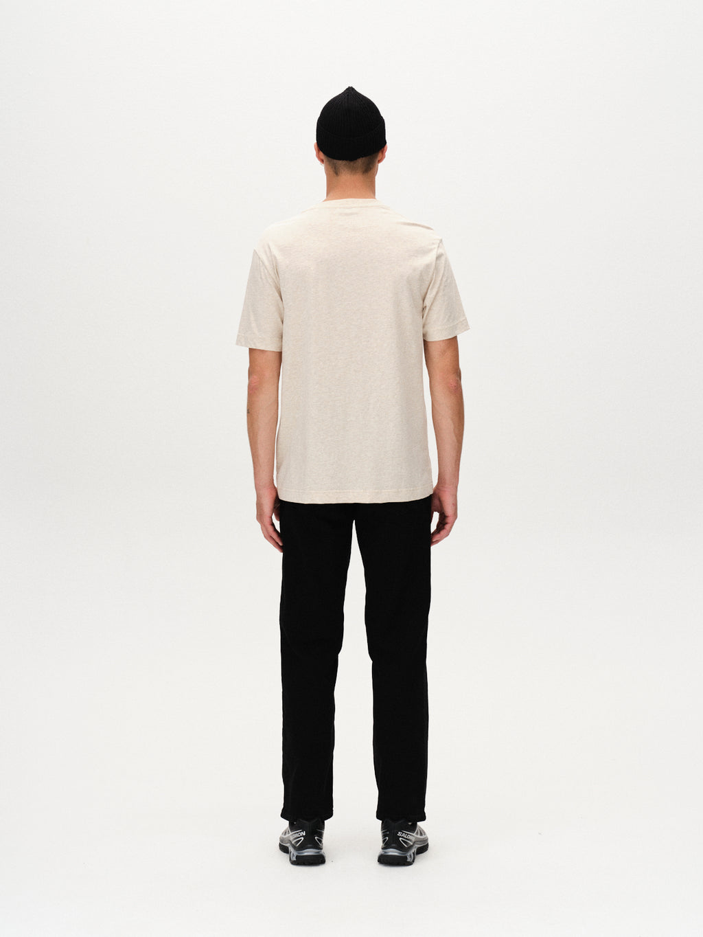 A person wearing the Gabba Dune Melange SS - Lt. Sand t-shirt, black pants, black shoes, and a black beanie stands facing away against a plain white background.