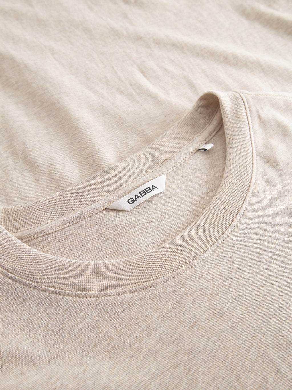 Close-up of the Dune Melange SS - Lt. Sand jersey T-shirt, made of ultra-soft fabric in a beige melange color. The Gabba logo is prominently displayed on the neckline tag. The shirt is laid flat to highlight its texture and weave.