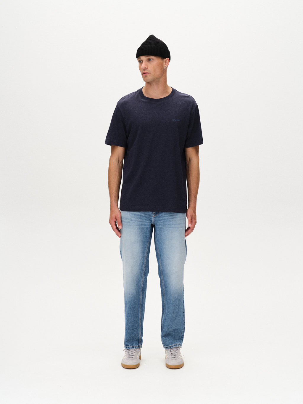 A man stands against a plain white background, looking slightly to his right, wearing a black beanie, Gabba Dune Melange SS - Navy Mel. t-shirt, light blue jeans, and white sneakers.