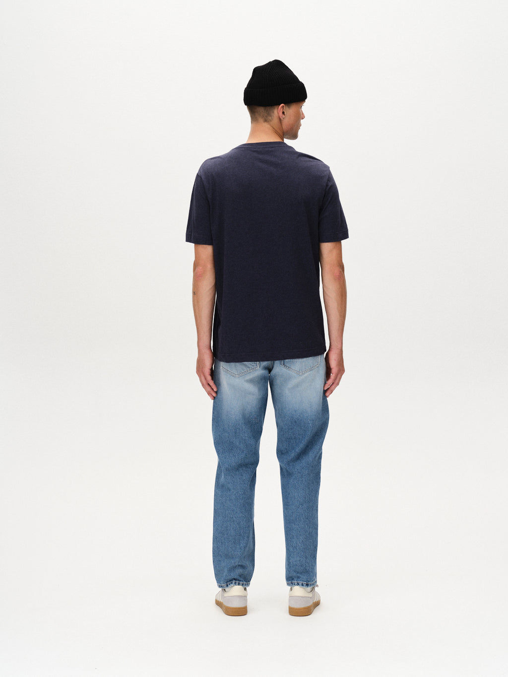 A person wearing a black beanie, light blue jeans, white sneakers, and a Gabba Dune Melange SS - Navy Mel. T-shirt stands facing away against a plain white background.
