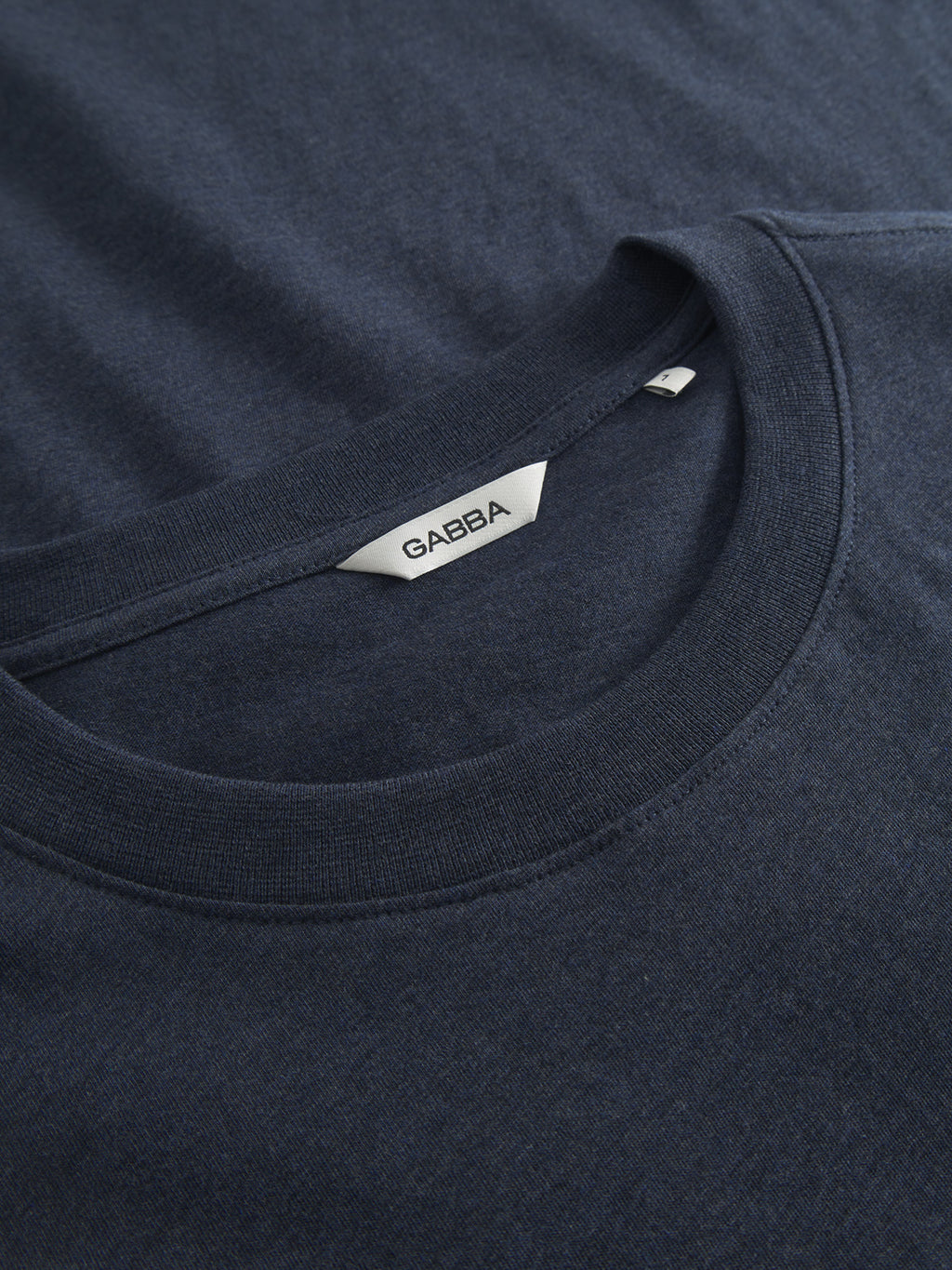 Close-up of the folded navy melange Dune Melange SS T-shirt by Gabba. The ultra-soft fabric resembles textured melange jersey with the Gabba logo tag and an inside-sewn label at the crew neckline.