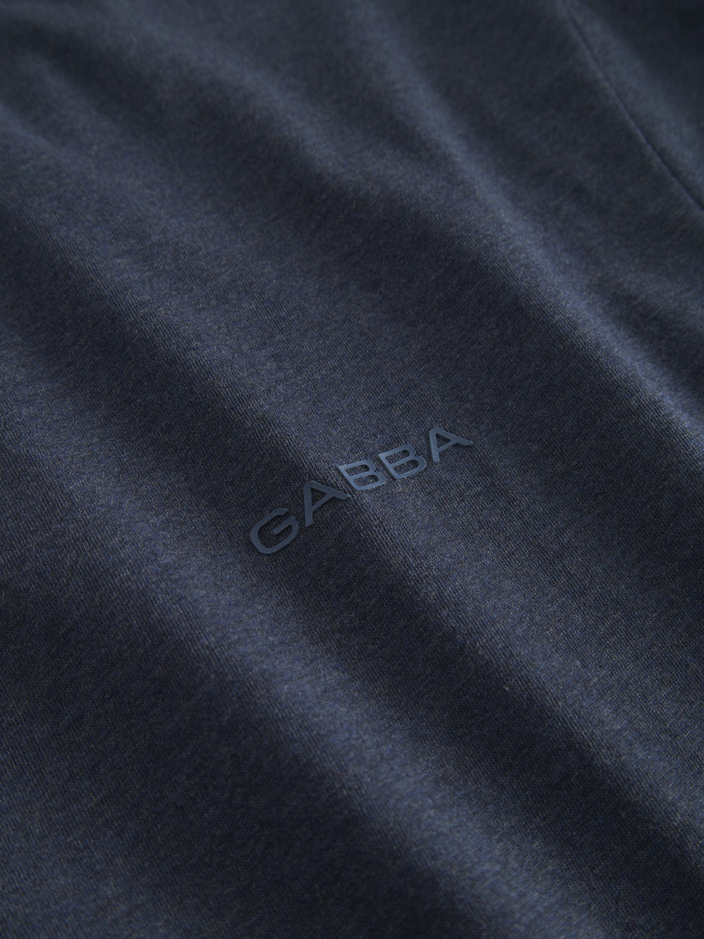 A close-up of the Gabba Dune Melange SS in Navy Mel reveals the jersey fabric with the Gabba logo embossed at its center, showcasing a smooth surface and subtle sheen for an ultra-soft texture.