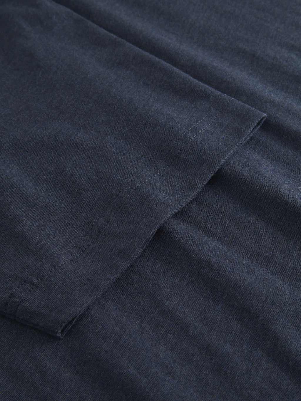 Close-up of the Dune Melange SS - Navy Mel. jersey by Gabba, featuring dark gray melange fabric with visible seam stitching. The ultra-soft, slightly textured material has folded edges overlapping on a flat surface.