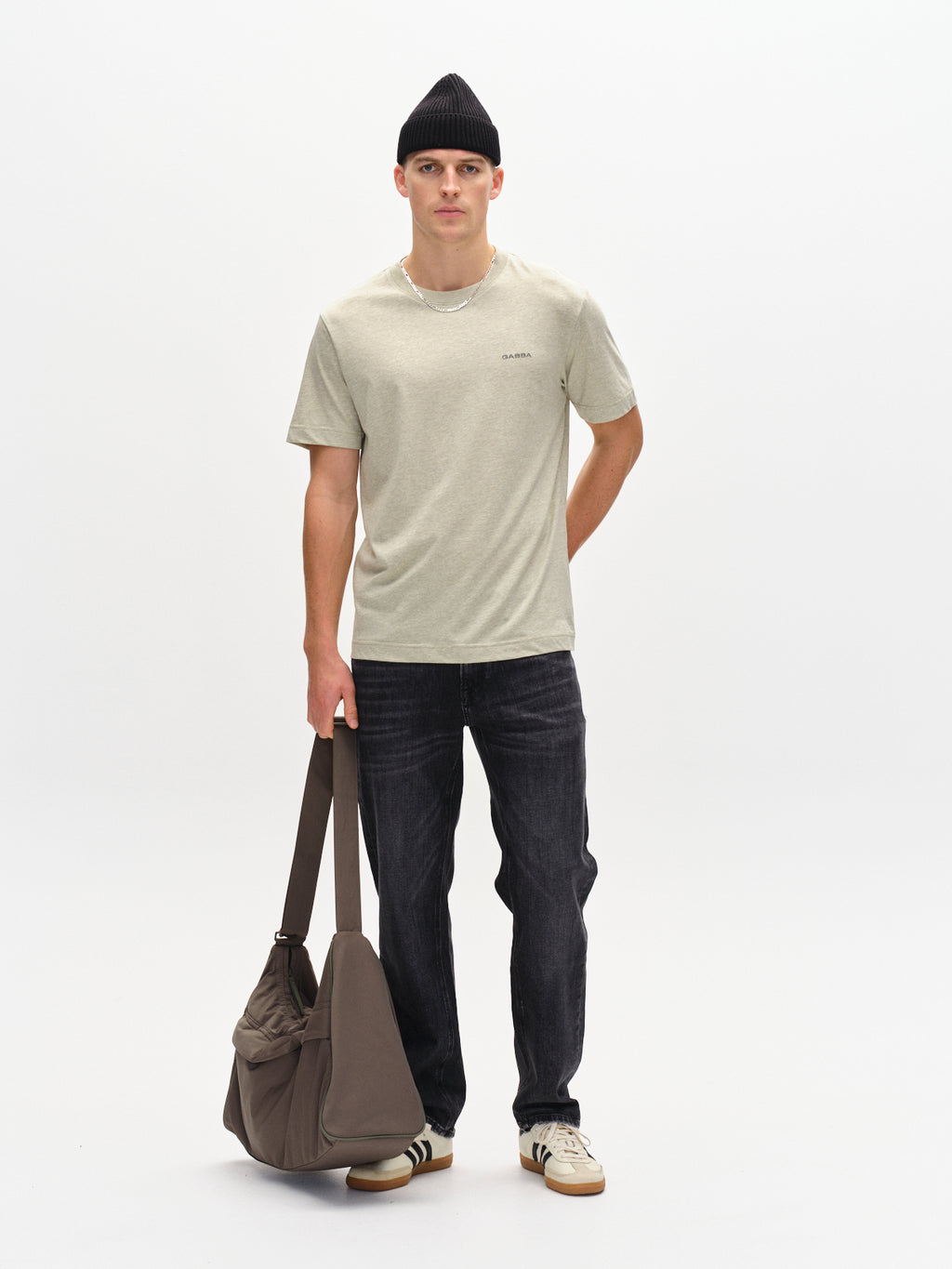 Against a plain backdrop, a casually dressed man in Gabbas green Dune Melange SS jersey T-shirt is paired with dark jeans and white sneakers. A black beanie completes his look, as he carries a brown duffel bag.