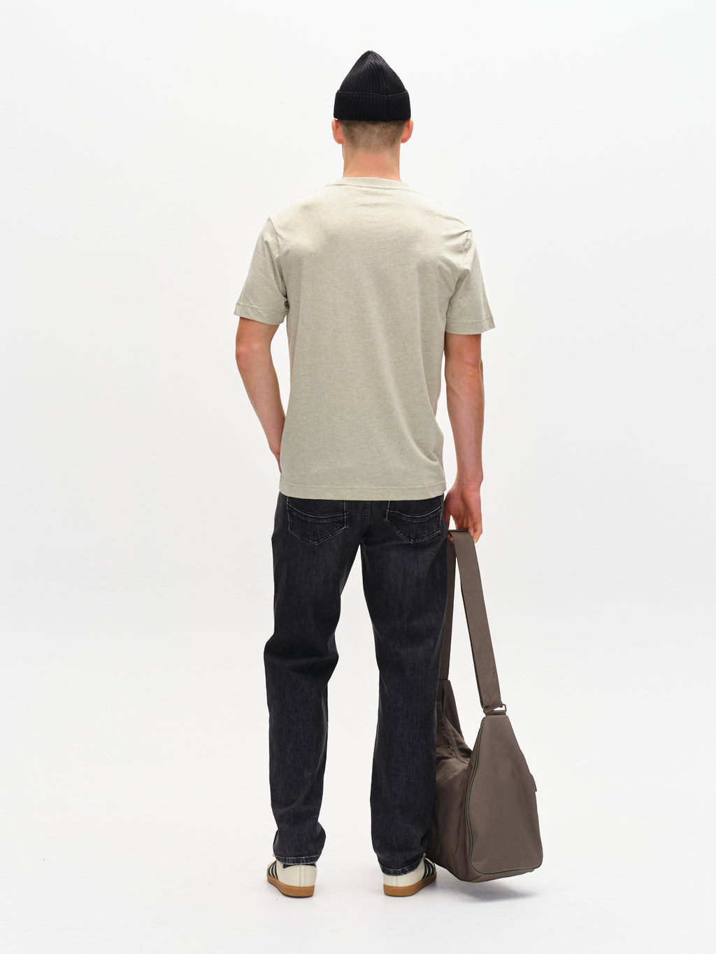 A person with a black beanie faces away, wearing Gabbas Dune Melange SS in Green Melange, dark jeans, and casual shoes. Theyre holding a large brown duffel bag against a plain white background.