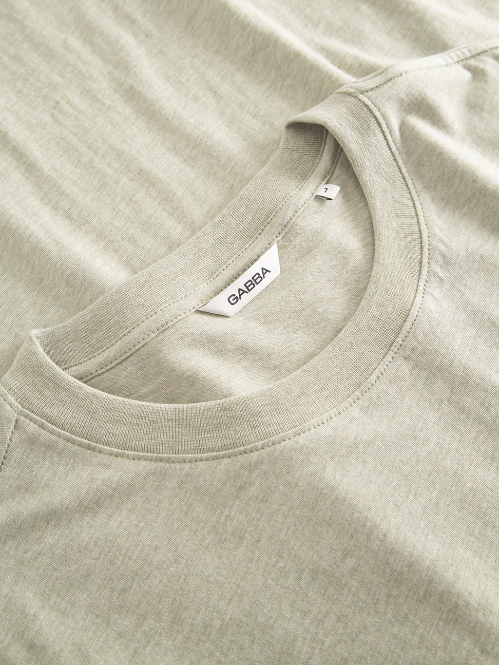 Close-up of the ultra-soft Dune Melange SS T-shirt in Green Melange by Gabba, featuring a visible neckline and logo label. Displayed flat, it highlights its smooth fabric texture and stitching details.
