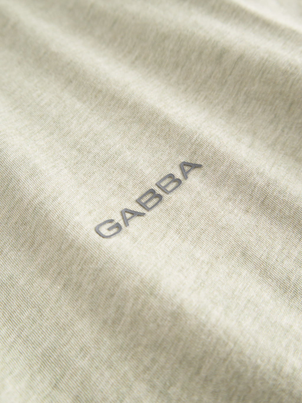 Close-up of the Gabba Dune Melange SS t-shirt in ultra-soft green melange jersey, showcasing a subtle texture with the logo printed in small, bold, dark gray letters at the center.