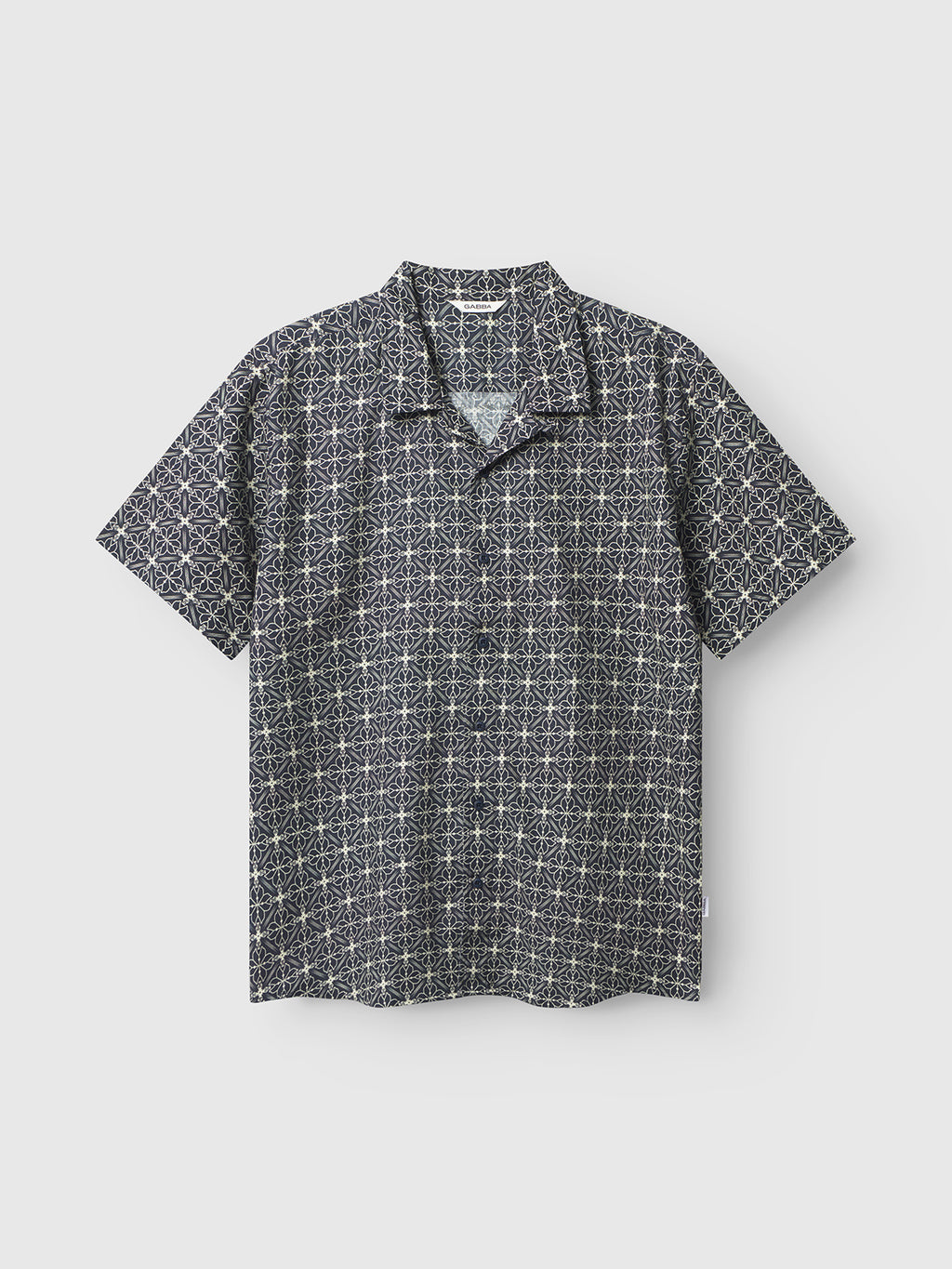 The Timo Resort AOP SS by Gabba is a premium short-sleeved, button-up shirt featuring an intricate white geometric star pattern on a navy background. Made from a linen-cotton blend, it includes a flat collar and is elegantly contrasted against a plain white backdrop.