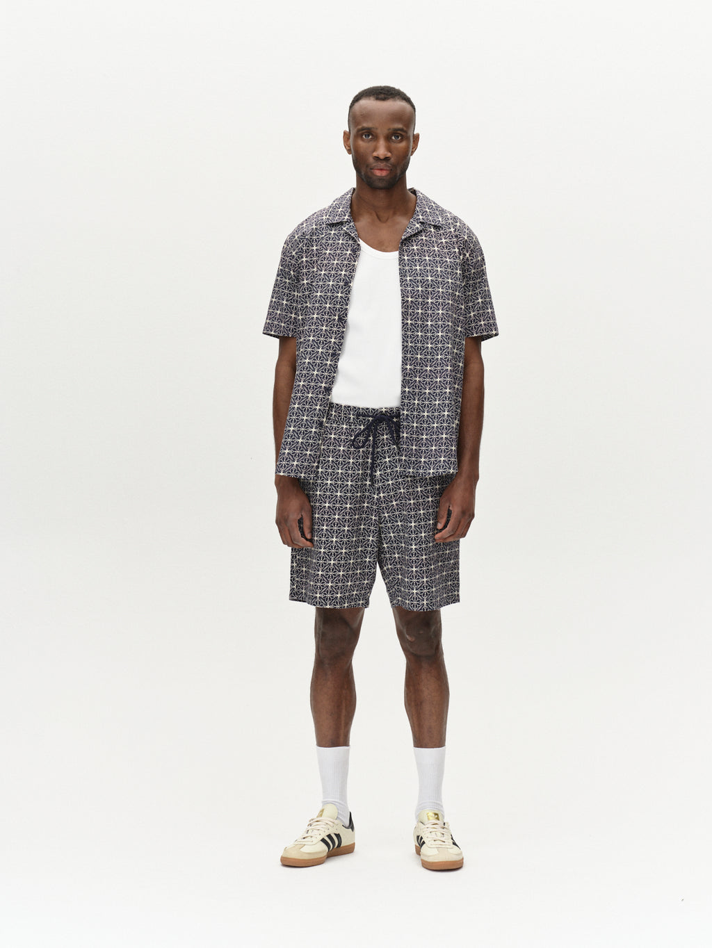 A person stands against a plain background wearing the Gabba Timo Resort AOP SS in navy and matching shorts over a white tank top, paired with white socks and beige sneakers featuring dark stripes, looking directly at the camera.
