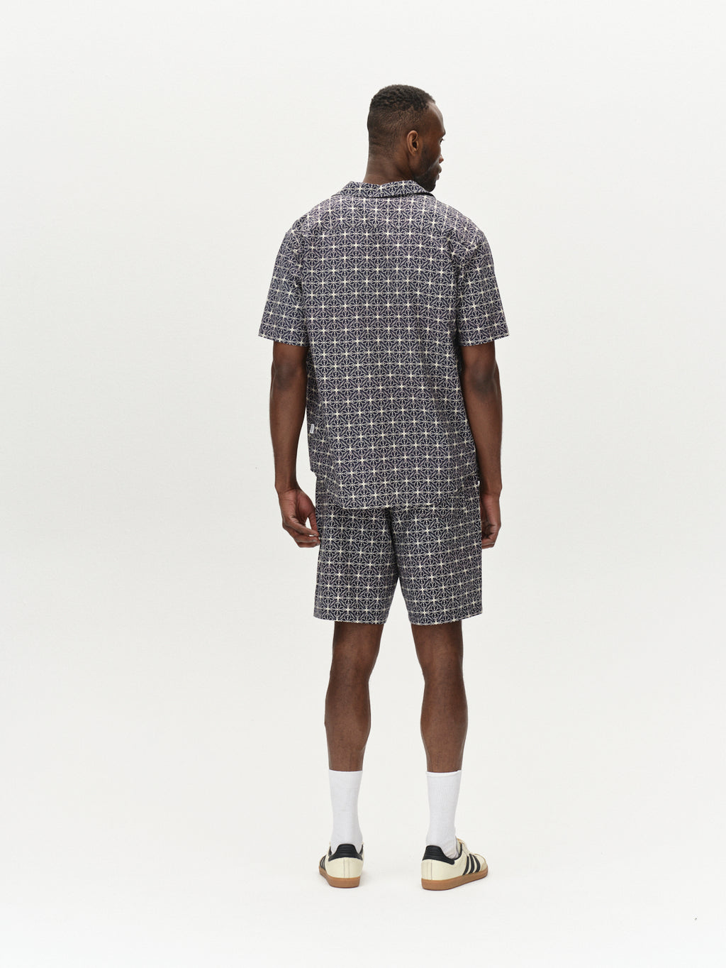 A man stands with his back turned, wearing the Timo Resort AOP SS - Navy shirt and shorts set by Gabba. His short hair complements his look, along with white socks and beige-black sneakers. The plain light-colored backdrop emphasizes the tasteful print of his outfit.
