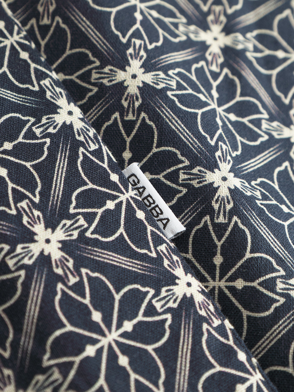A close-up shows a seasonal geometric floral pattern in white on navy from the Timo Resort AOP SS by Gabba. The luxurious linen-cotton blend fabric features a small label reading Gabba, ideal for crafting a premium shirt.