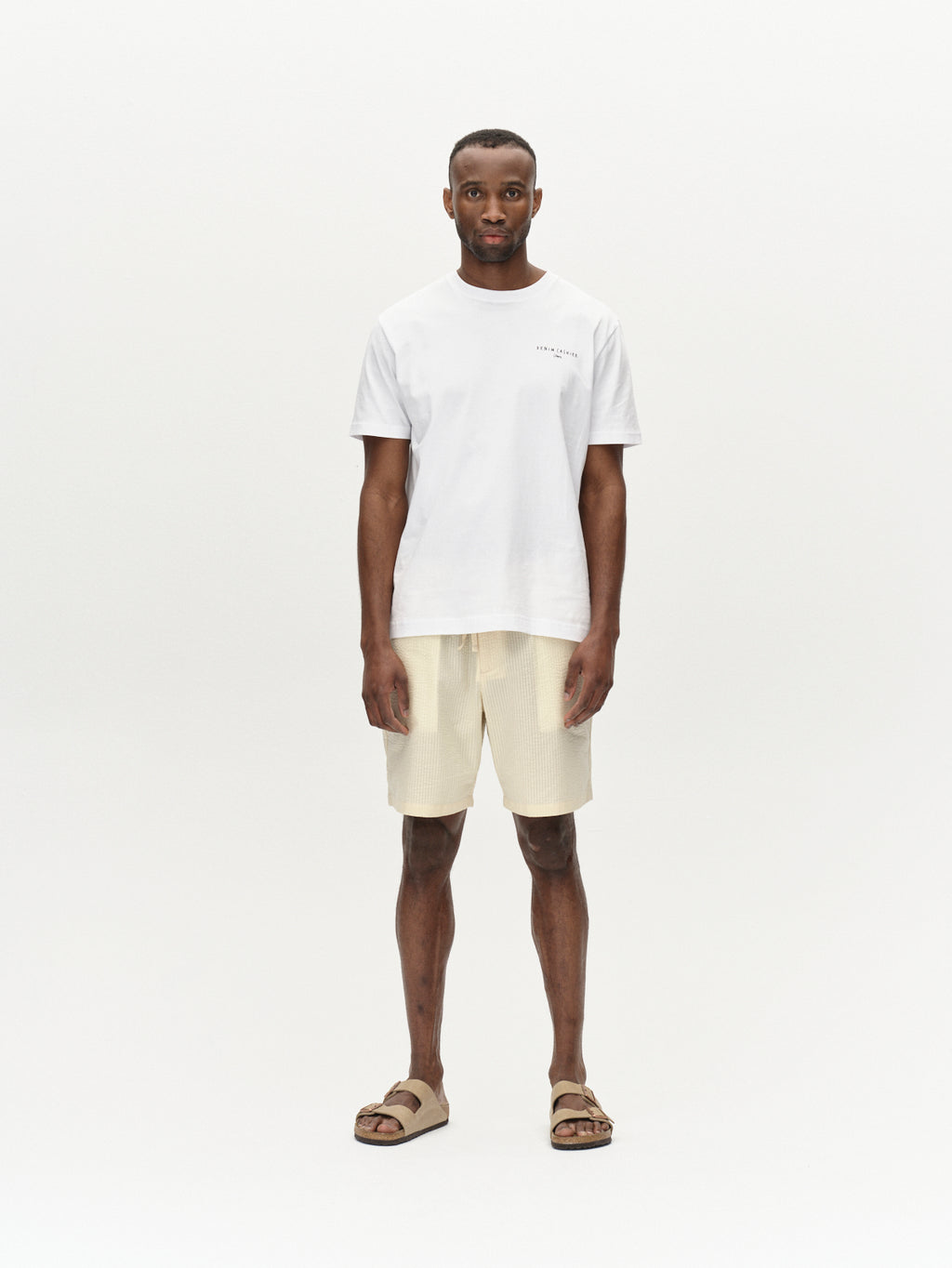 A man stands against a plain background, wearing a white t-shirt with Gabbas Tommi Seer - Ecru Light premium seersucker shorts and beige sandals. His arms relax by his sides, exuding effortless style.