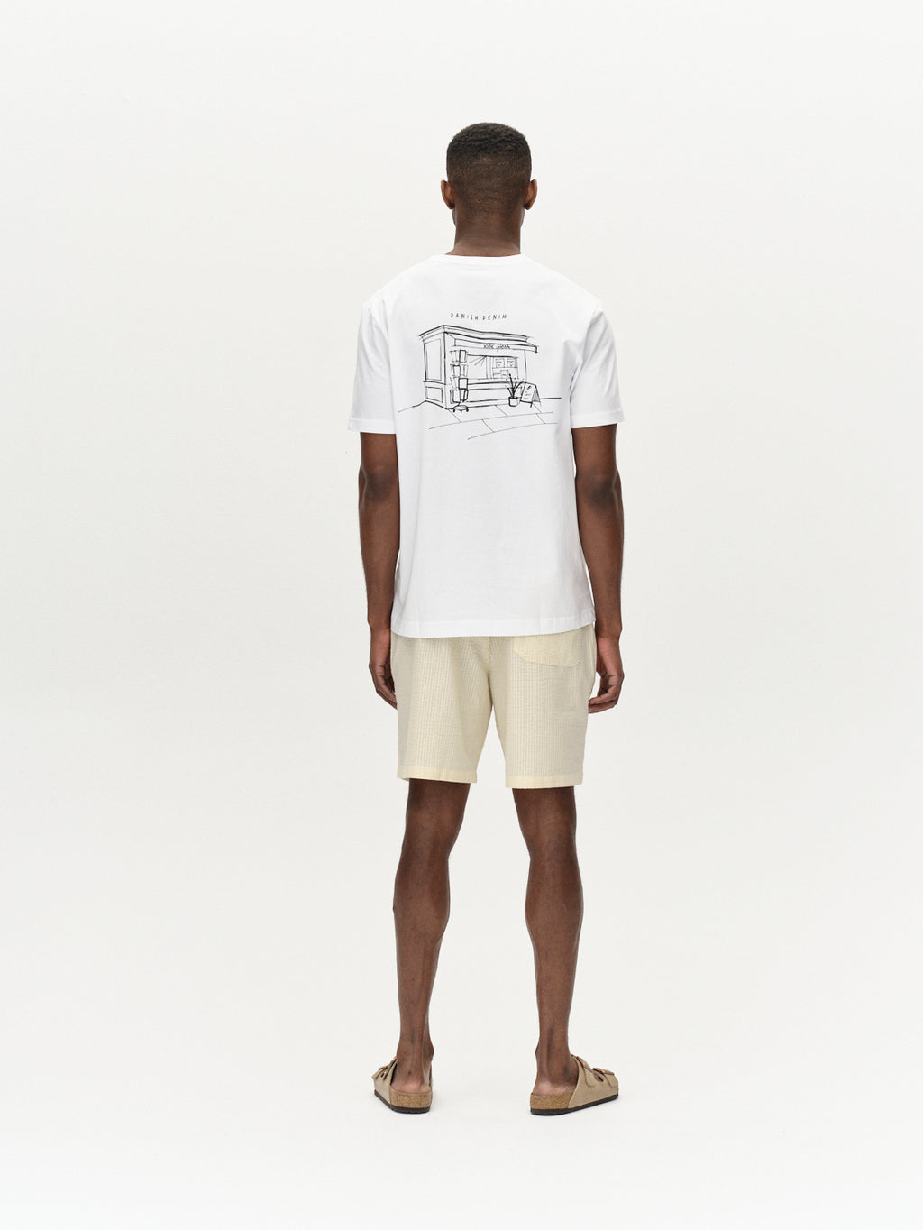 A person stands against a plain white background, facing away, wearing a Gabba Tommi Seer - Ecru Light t-shirt with a minimalist house scene line drawing and text on the back, paired with beige premium shorts and sandals.