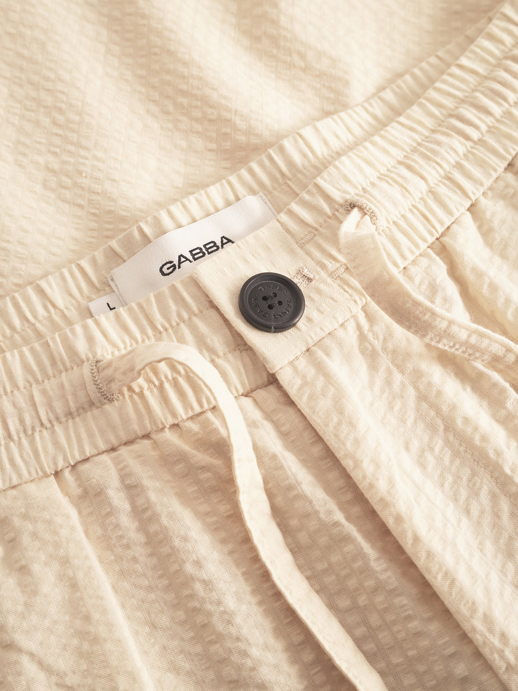 Close-up of Gabbas Tommi Seer - Ecru Light beige pants made from 100% cotton seersucker fabric, featuring an elastic waistband with a drawstring and a dark button. A white label with the brand name Gabba is sewn inside the waistband.