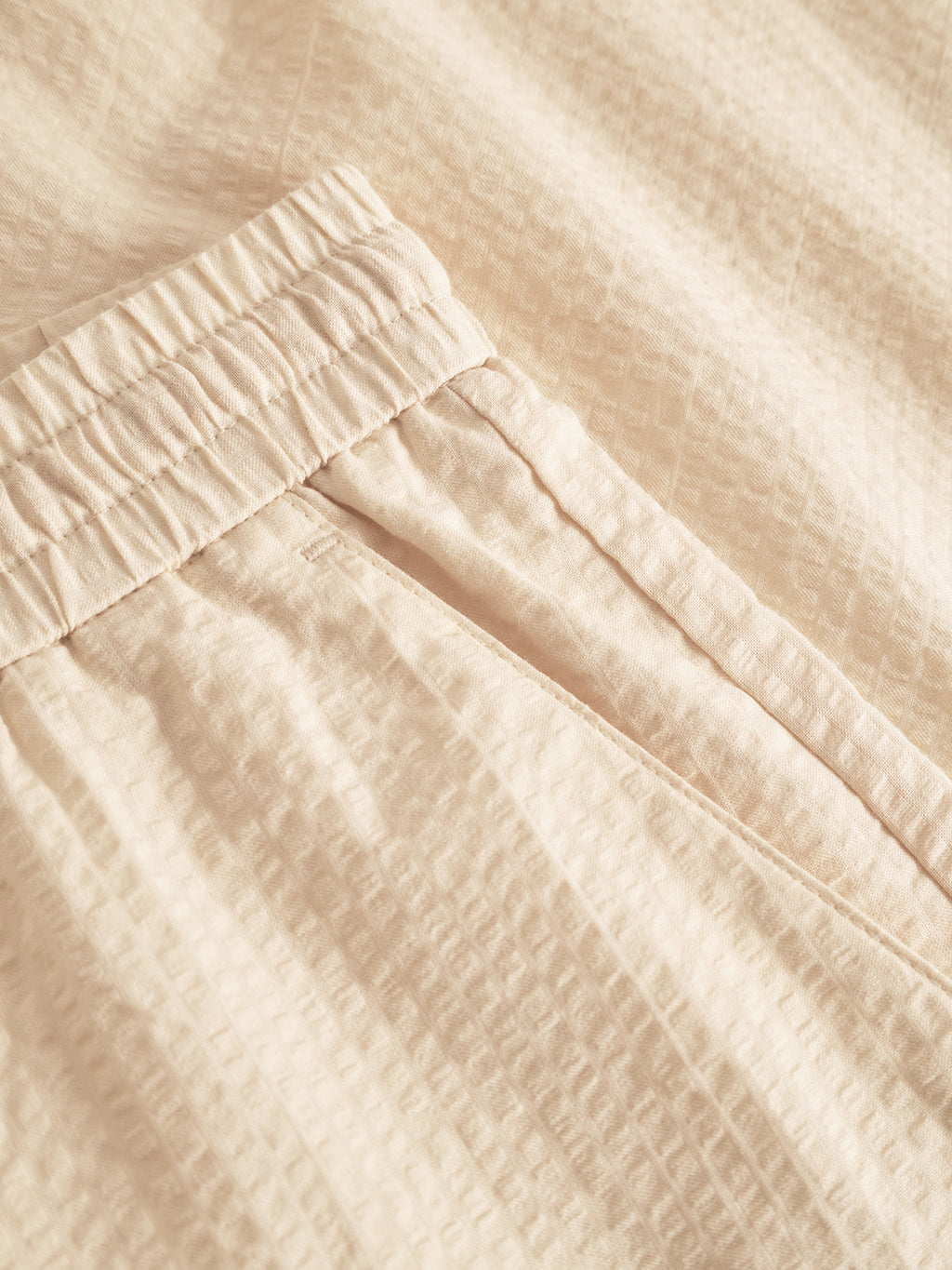 Close-up of Tommi Seer - Ecru Light pants by Gabba, featuring an elastic waistband and a side pocket. Crafted from 100% cotton, the fabric has a subtle textured pattern similar to seersucker, with detailed stitching visible in the image.