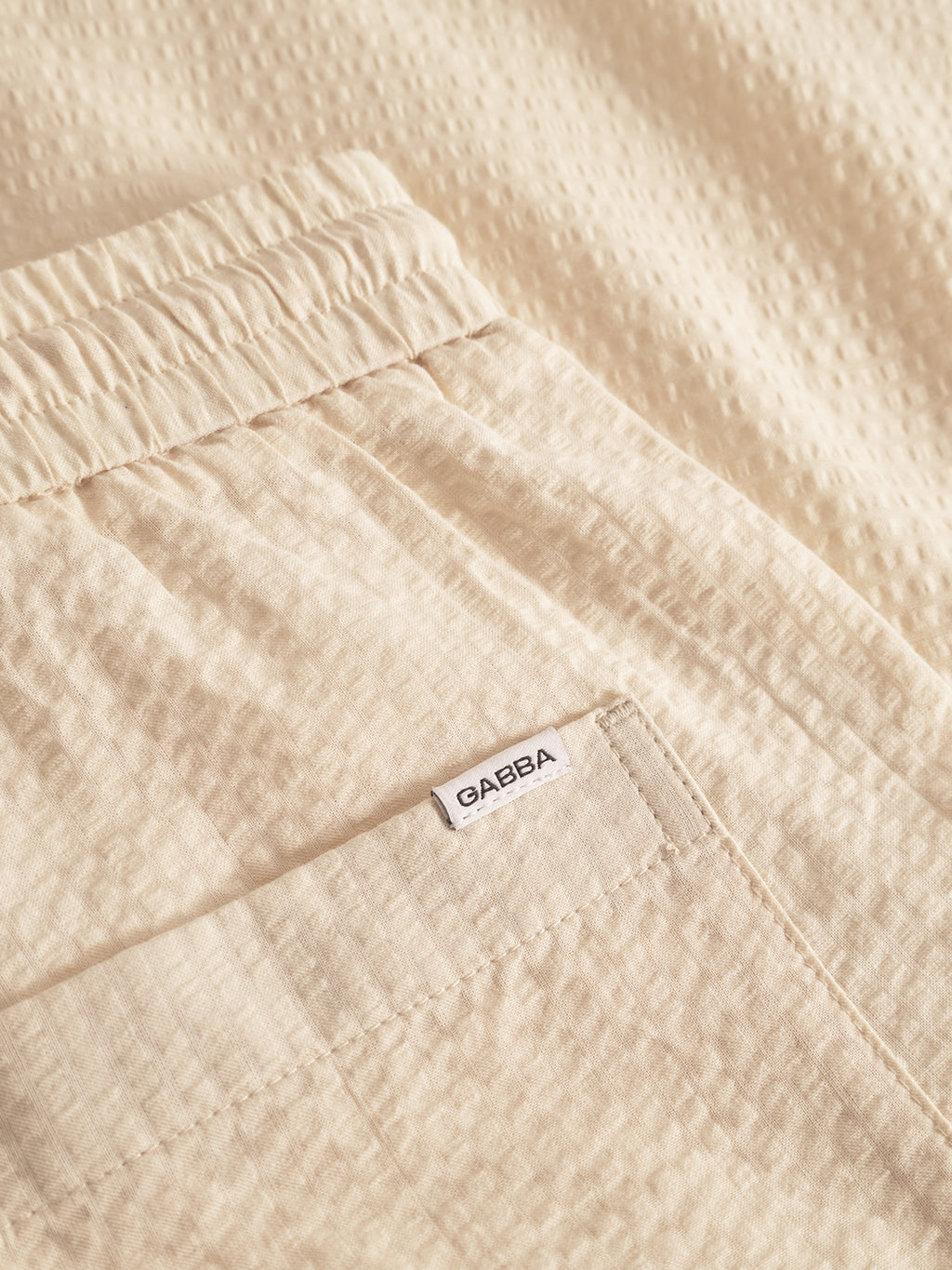 Close-up of Tommi Seer - Ecru Light shorts by Gabba in beige textured seersucker fabric. They feature an elastic waistband, a pocket with a small white Gabba label, and are crafted from 100% cotton for a subtle dotted pattern.