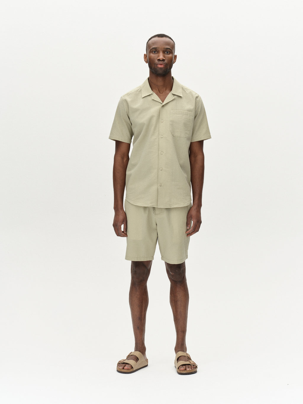 A man in a Gabba Tommi Seer Green Sea shirt and premium shorts stands against a plain white background, wearing beige sandals.