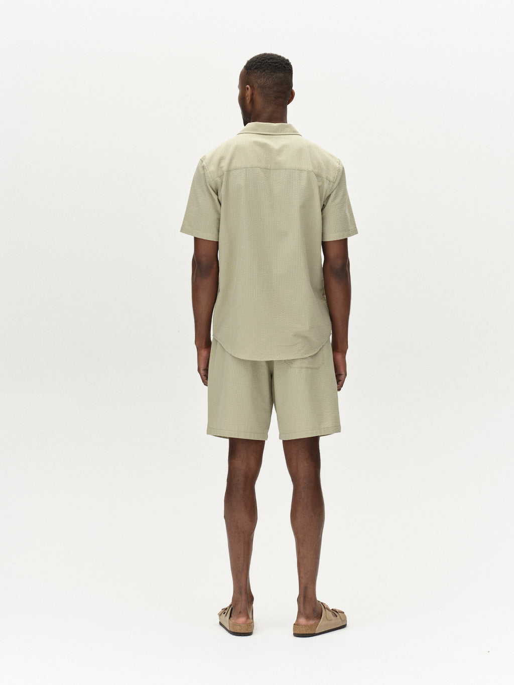 A person stands against a plain background, facing away, wearing Gabbas Tommi Seer - Green Sea shirt and premium seersucker shorts. The 100% cotton outfit features short sleeves and a monochromatic design, paired with sandals for a casual look.