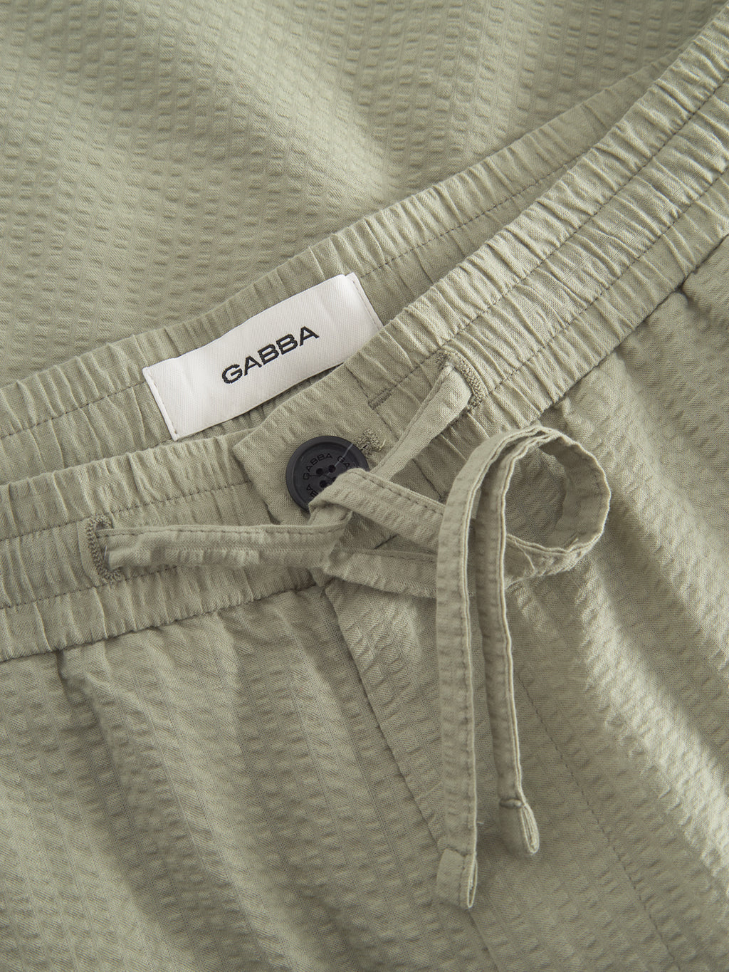 Close-up of Tommi Seer - Green Sea shorts by Gabba in light olive green seersucker fabric with a waffle texture, featuring an elastic waistband, visible drawstring, and a black button. A white GABBA label is sewn inside the 100% cotton material.
