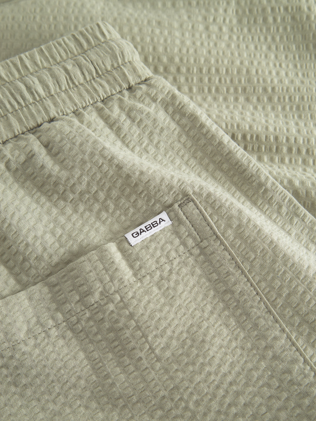 Close-up of Gabbas Tommi Seer - Green Sea shorts in light olive green textured fabric. Made from 100% cotton, they feature an elastic waistband and a small white tag with OAREBA near the pocket.