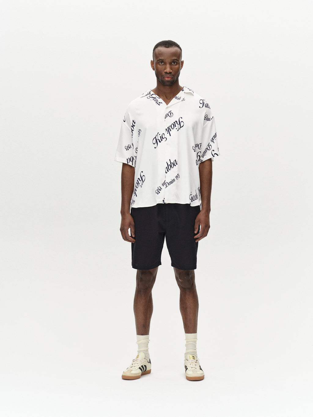 Standing against a white background, the man wears the Tommi Seer - Black ensemble by Gabba, featuring a 100% cotton white shirt with black cursive text and black shorts. He completes his look with short hair, white sneakers with black stripes, and beige socks for subtle charm.