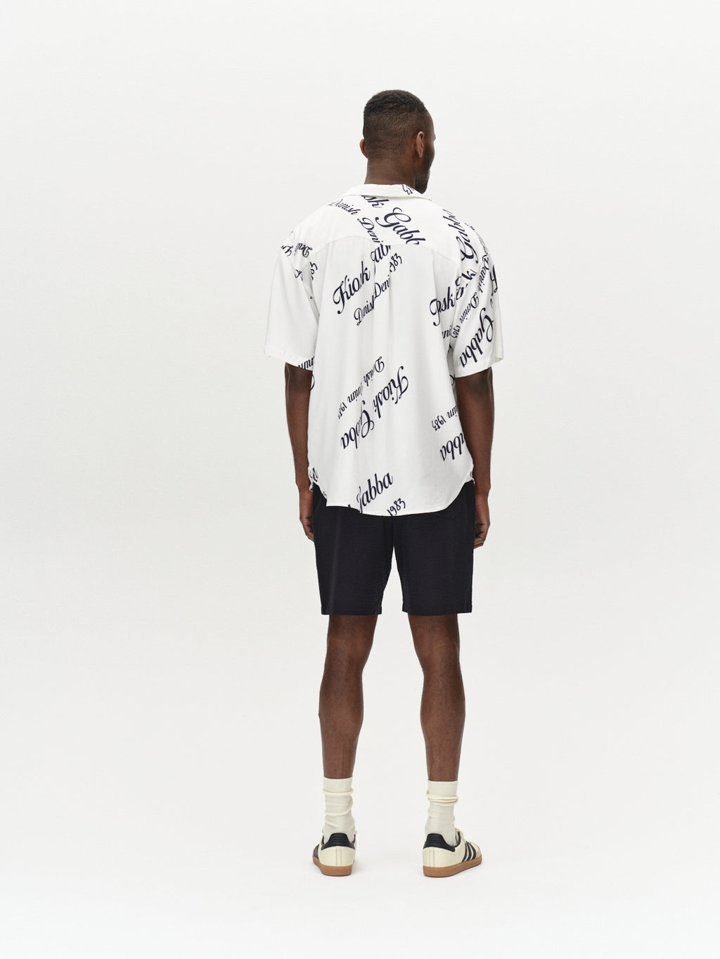 A man stands facing away in a white shirt with cursive text, paired with Gabbas Tommi Seer - Black premium black seersucker shorts, white crew socks, and sandals. A plain white background accentuates his effortlessly cool style.