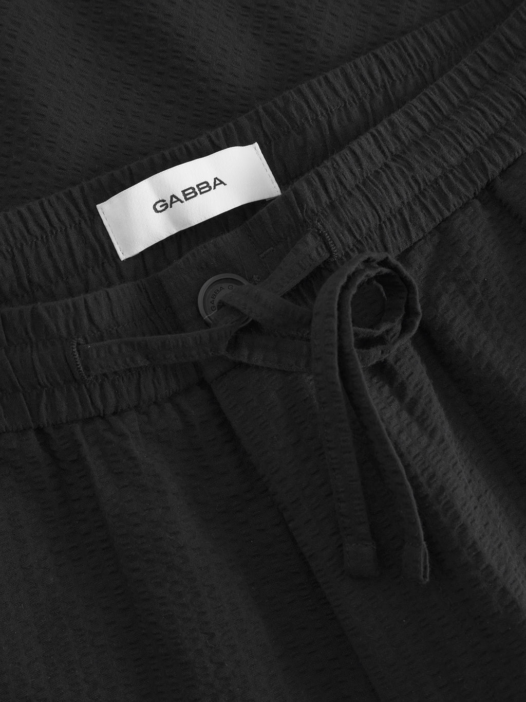 Close-up of Tommi Seer - Black pants crafted from seersucker, featuring an elastic waistband, drawstring, and button. A white Gabba label is inside.
