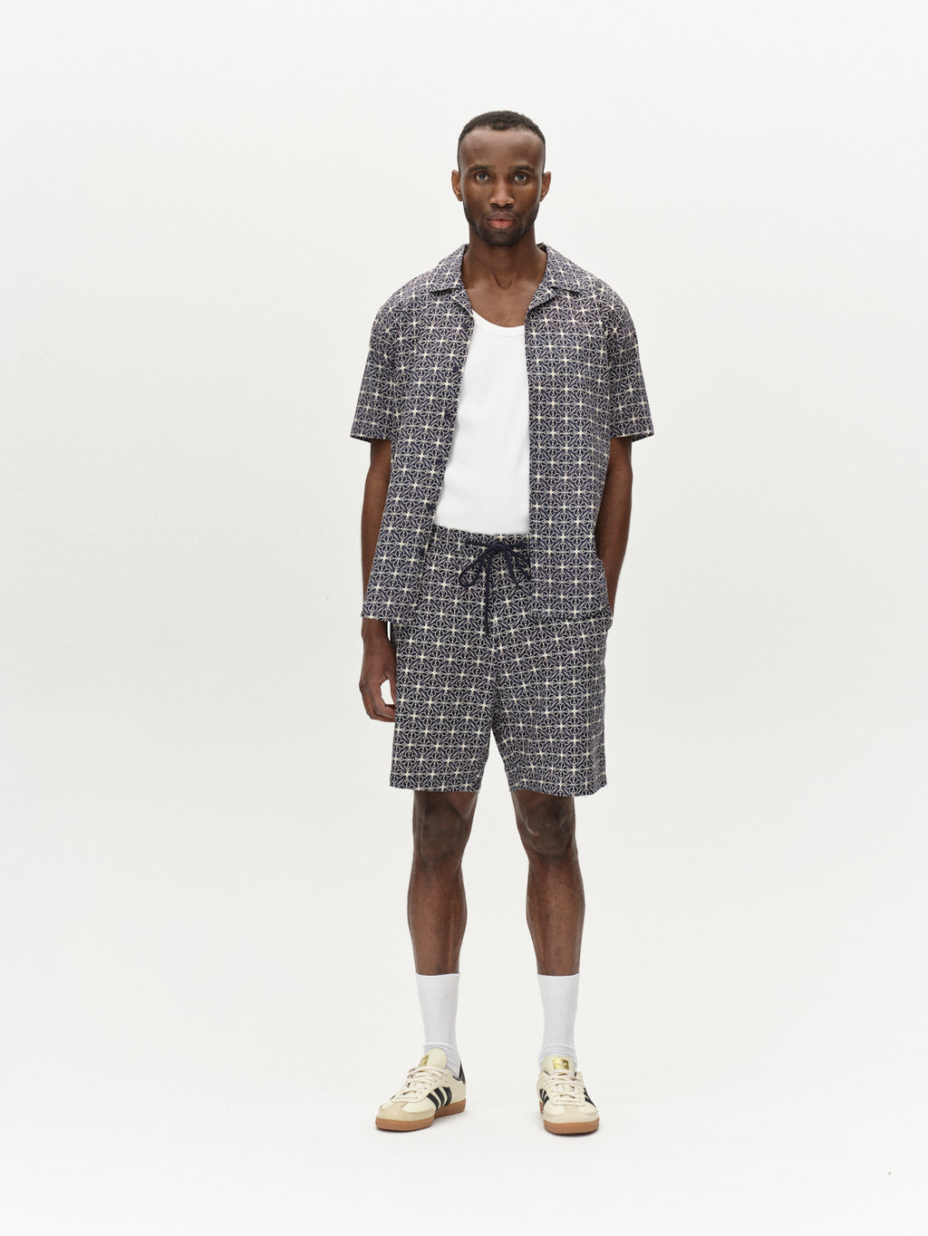 Against a white backdrop, a person wears the Gabba Tommi AOP in navy—a patterned seersucker short-sleeve shirt with matching shorts—paired with a white cotton tank top, white socks, and sneakers. With hands in pockets, they gaze forward effortlessly.