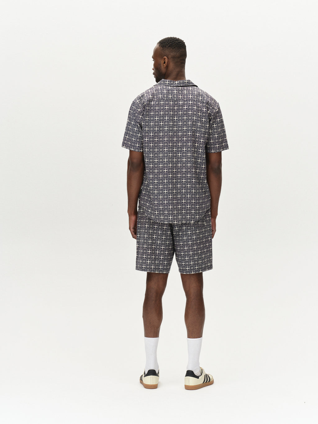 A man stands facing away in a matching short-sleeve shirt and shorts from Gabbas Tommi AOP - Navy. The outfit, featuring a black and white geometric pattern, is crafted from soft cotton. He sports short hair, white socks, and striped sneakers against a plain white background.