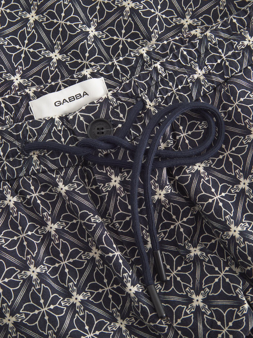 A close-up of Tommi AOP - Navy by Gabba shows intricate white floral patterns on a dark background, resembling seersucker fabric. It includes a white GABBA label and dark drawstrings with black tips, indicating it is part of a stylish pair of cotton shorts.