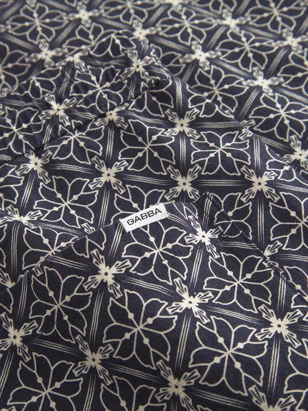 Close-up of navy seersucker fabric with a geometric floral pattern in white. A small tag reading Gabba is sewn onto the cotton, ideal for stylish Tommi AOP shorts.
