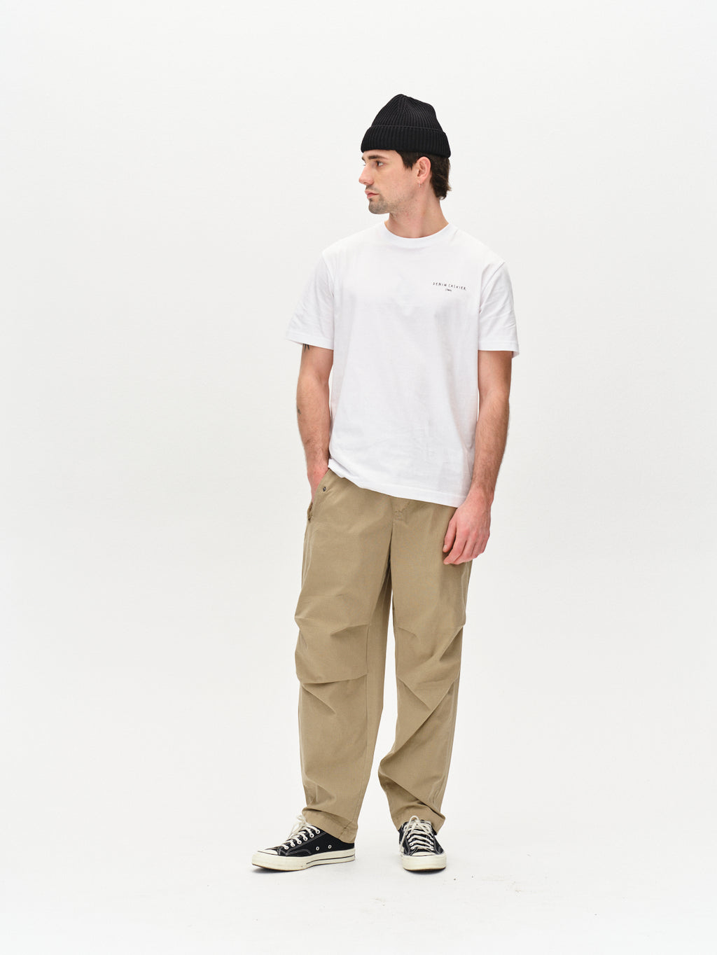 A person dons a white t-shirt, Gabbas Topgun Jash - Sand beige technical pants with an elastic waistband, a black beanie, and black sneakers with white soles. They stand against a plain white background, hands in pockets, gazing sideways.