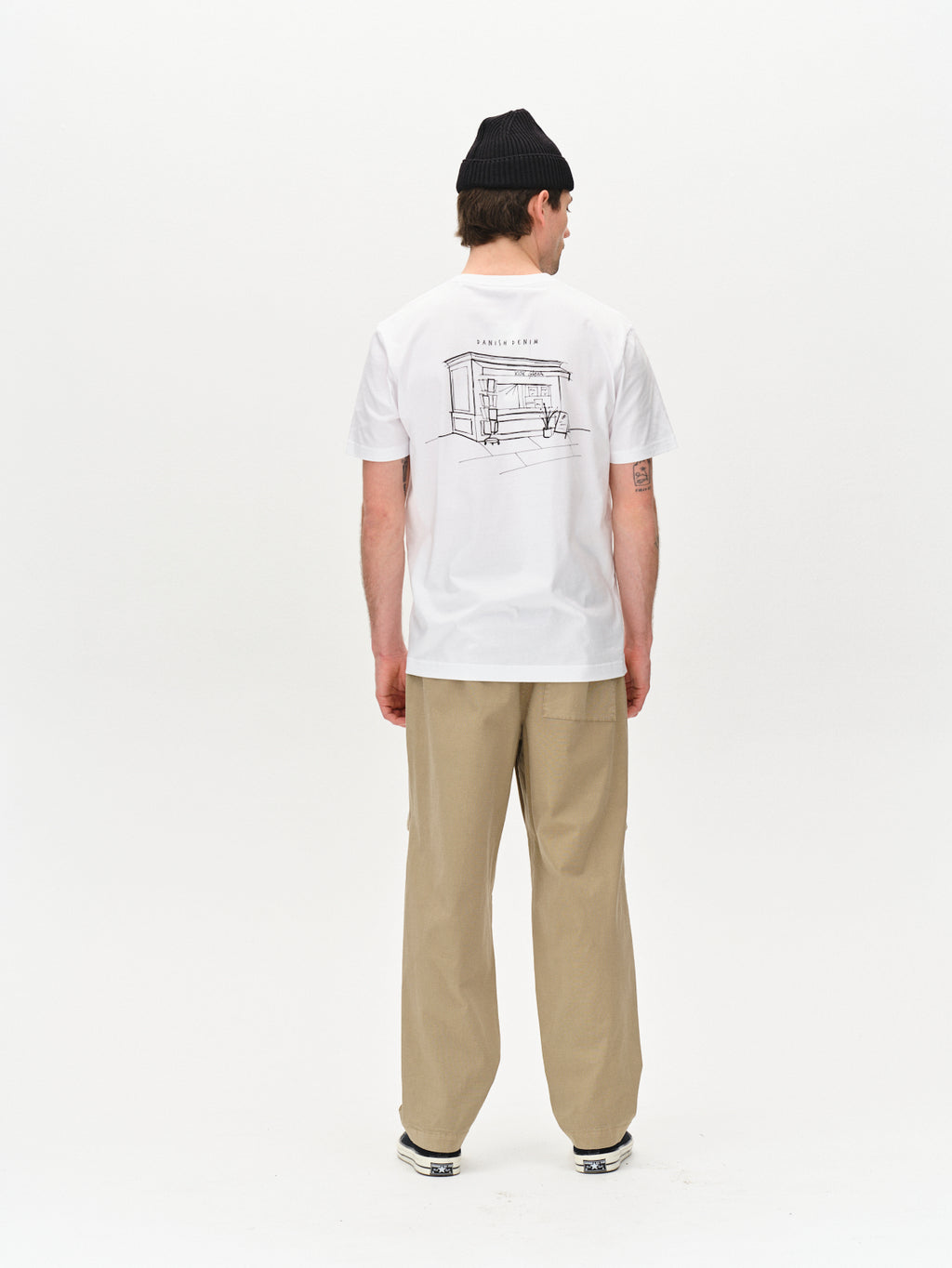 A person wearing a black beanie, the Gabba white graphic t-shirt featuring a house sketch with House Seiji on the back, Topgun Jash - Sand beige trousers with an elastic waistband, and sneakers stands facing away against a plain white background.