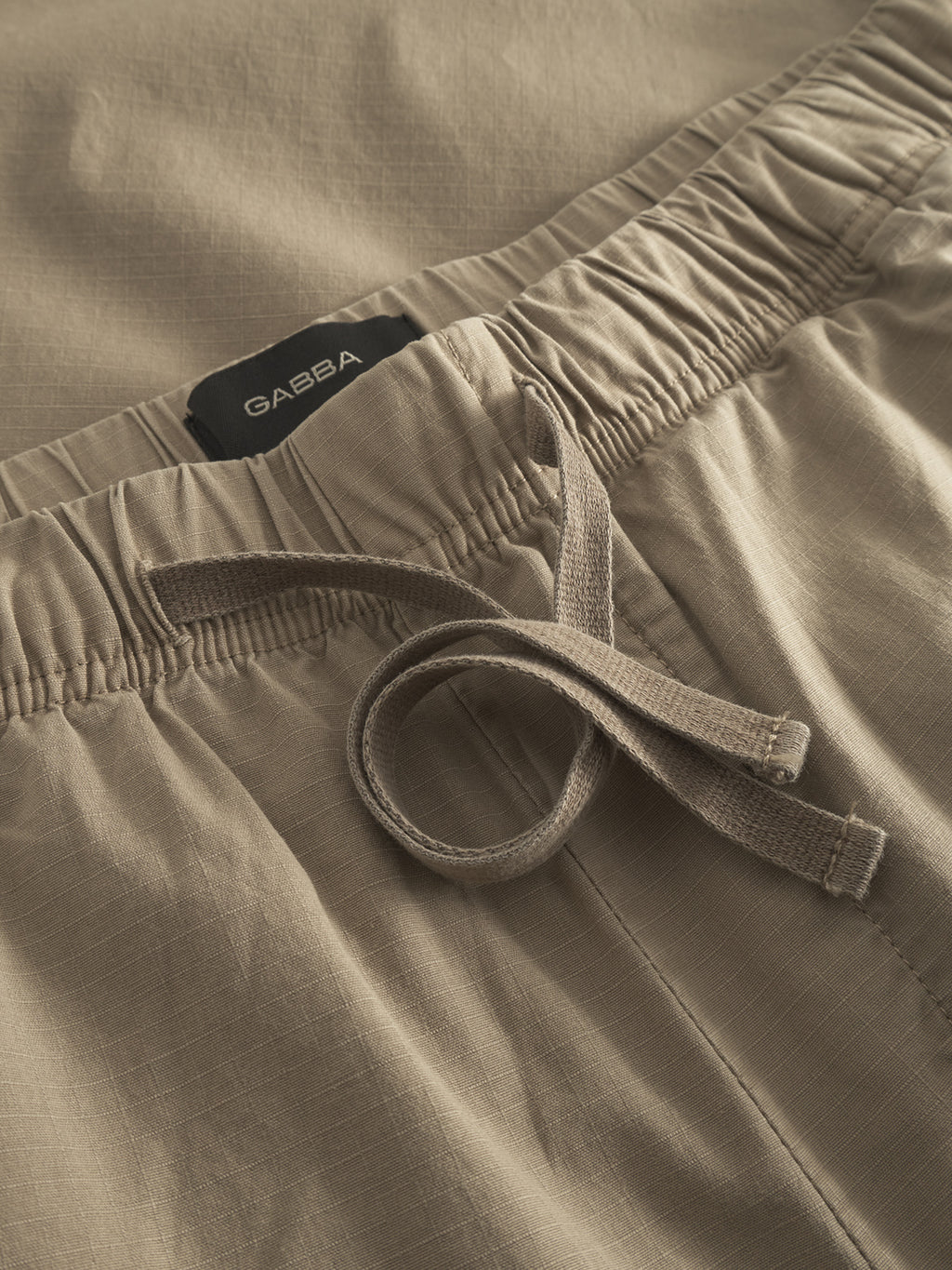 Close-up of the Topgun Jash - Sand pants by Gabba, highlighted by an elastic waistband with a drawstring, crafted from soft ripstop fabric with a subtle grid pattern and accented by a black label between the waistband and fabric.