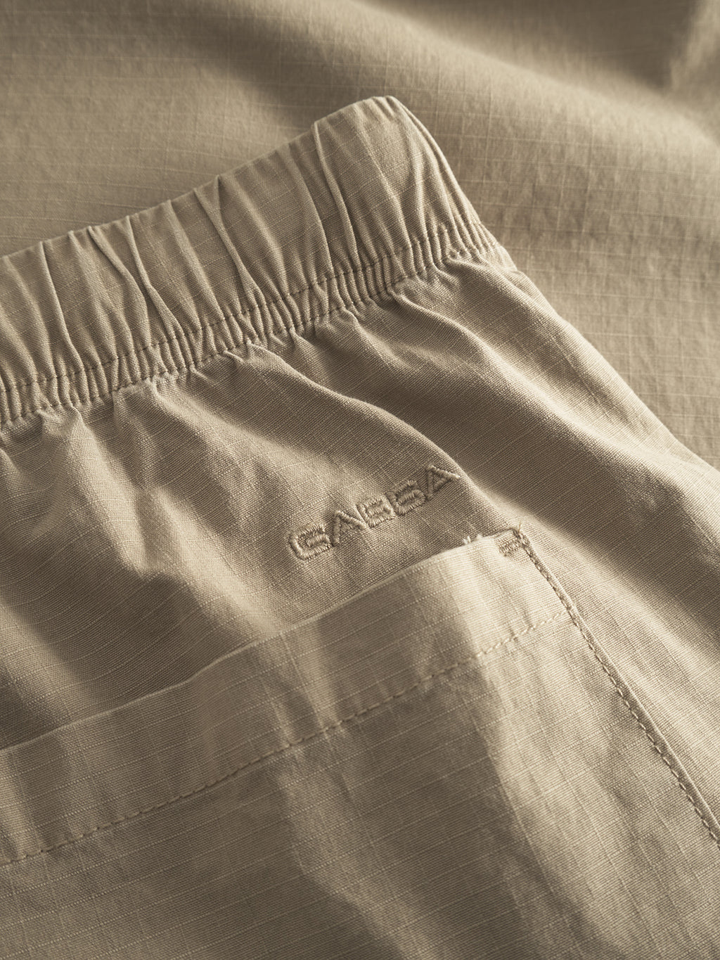 A close-up of the Topgun Jash - Sand pants highlights the smooth, slightly crinkled texture of the lightweight ripstop fabric. Featuring an elastic waistband and pocket seam, Gabba is subtly embroidered near the waist.