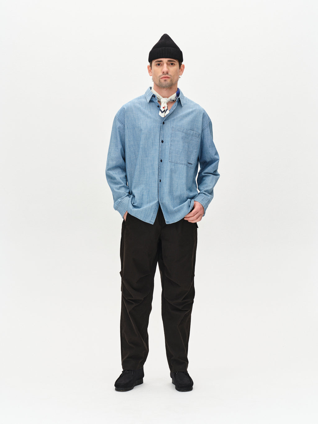 A person stands against a plain white background wearing a blue button-up shirt, Gabbas Topgun Jash black technical pants with an elastic waistband, a black beanie, and black shoes. They have a tattoo on their neck and hands in pockets, exuding a casual style.