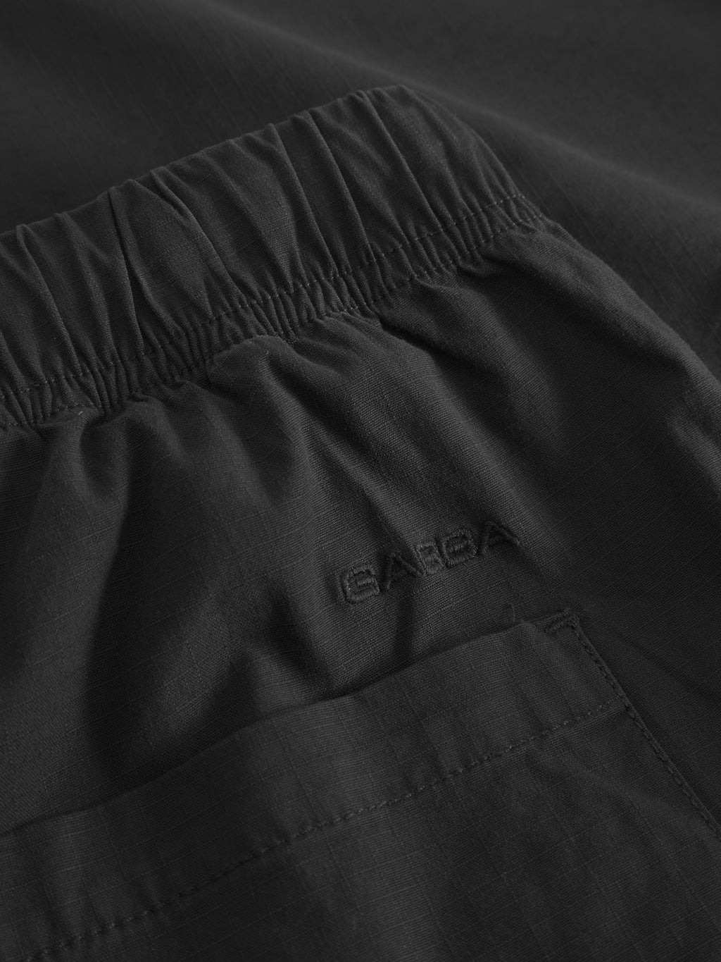 Close-up of Gabbas Topgun Jash pants in black, featuring an elastic waistband and back pocket. The ripstop fabric has a subtle grid texture, and the brand name is embroidered in dark stitching above the pocket.