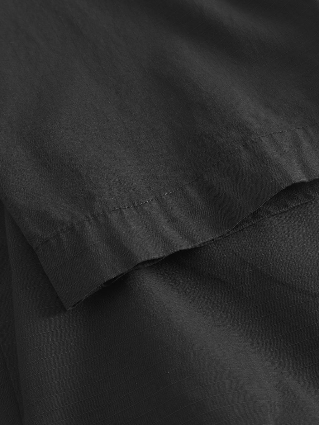 Close-up of overlapping layers of Topgun Jash - Black by Gabba, showcasing dark gray ripstop fabric with visible seams and subtle stitching. The material looks soft and slightly crumpled, forming gentle shadows.
