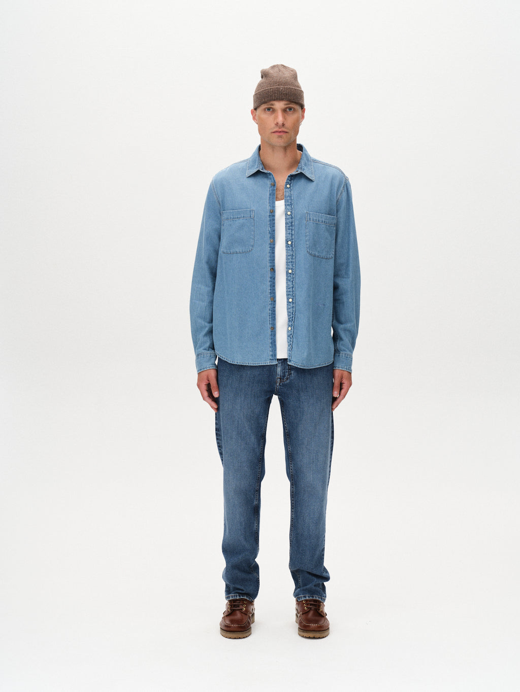 A man stands against a white background, wearing the Gabba Sven Reg Denim LS shirt in light blue denim with chest pockets over a white t-shirt, blue jeans, brown shoes, and a beige knit beanie for a vintage-inspired look.