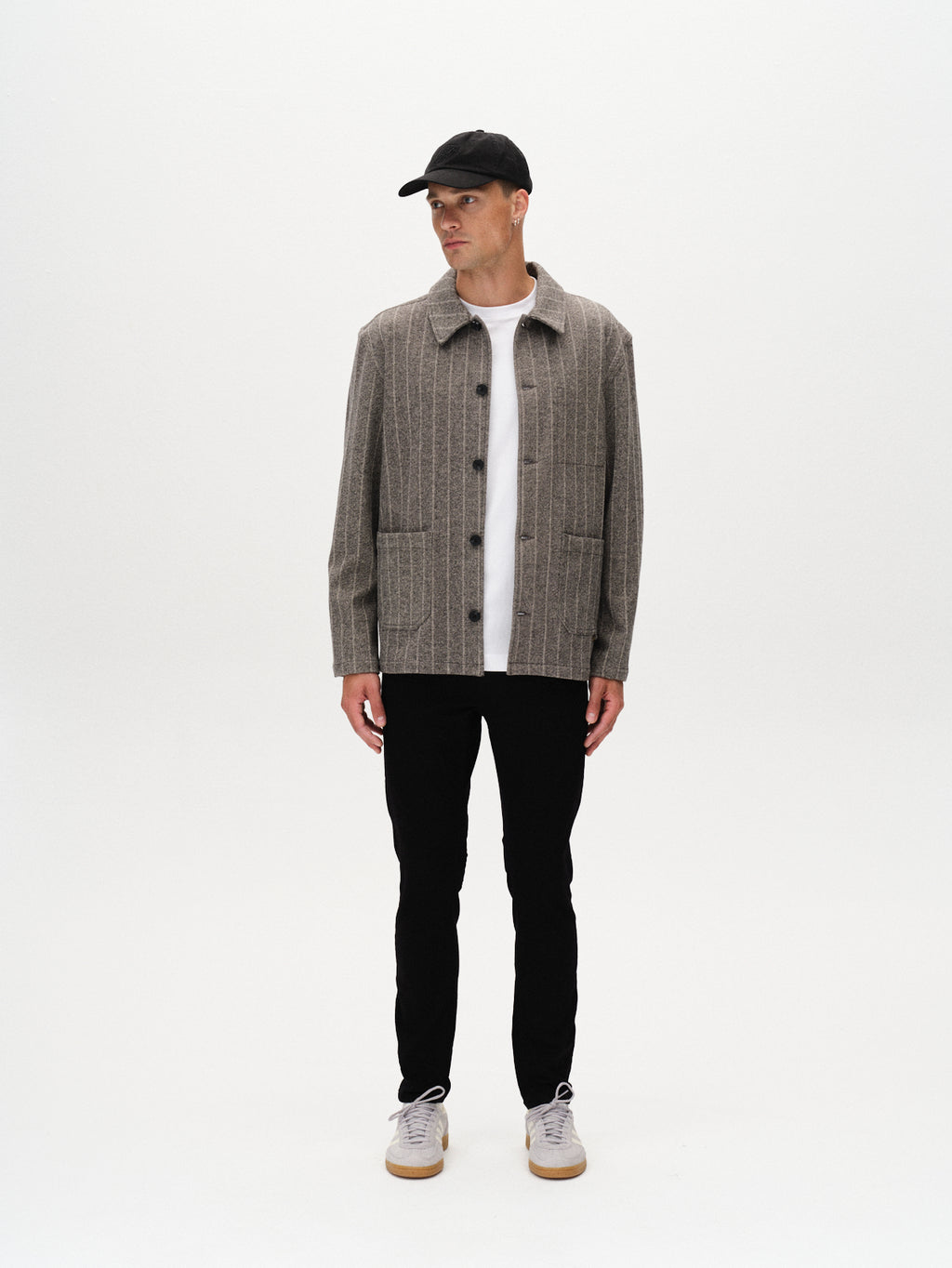 A man wearing Gabbas Jones Hold Black - Black Denim jeans, a black cap, gray striped button-up jacket, white T-shirt, and white sneakers stands against a plain white background, looking left with arms relaxed at his sides.