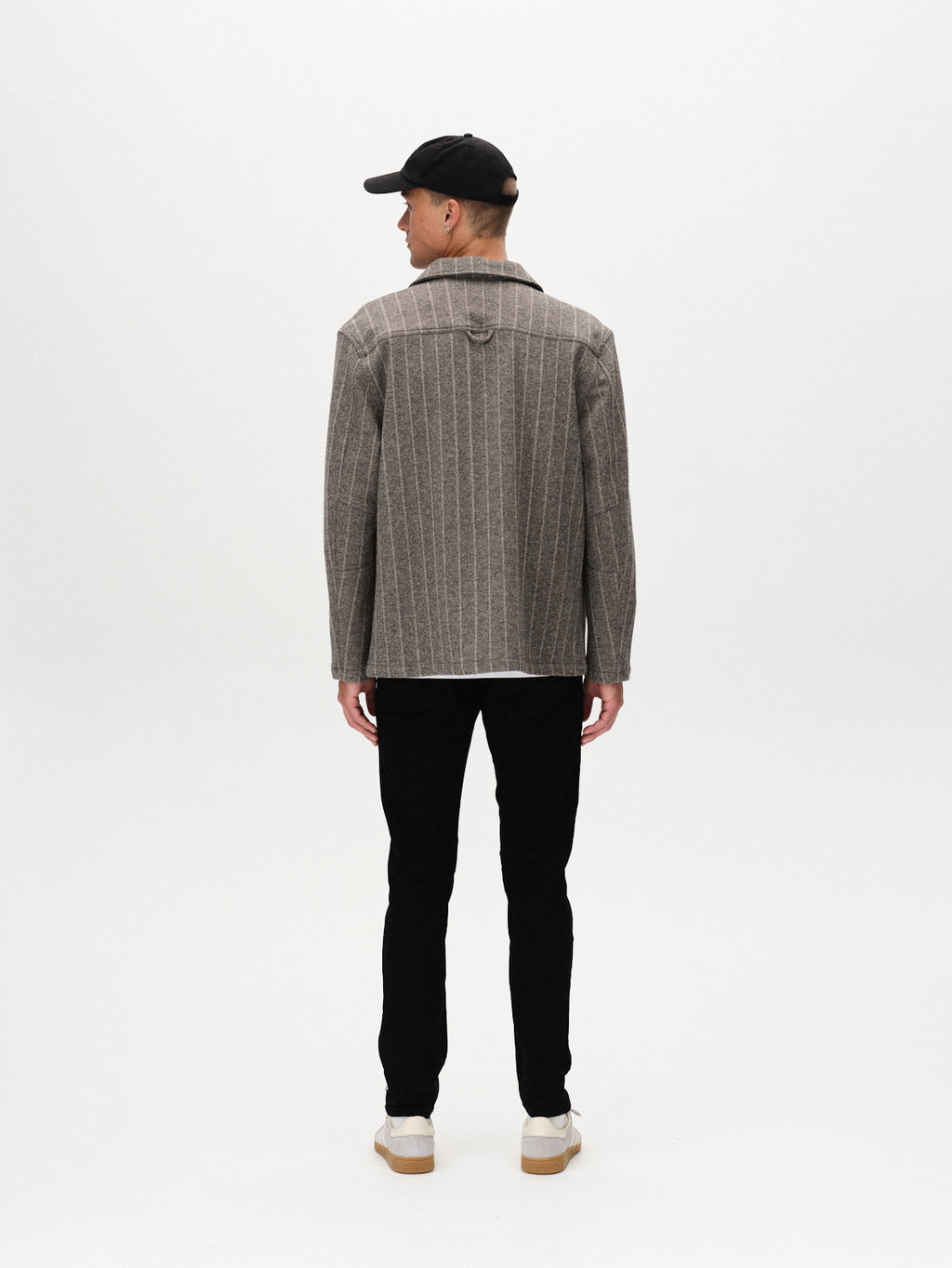 A man stands facing away from the camera, wearing a black cap, gray pinstriped jacket, white sneakers, and Gabba Jones Hold Black - Black Denim jeans against a plain white background.