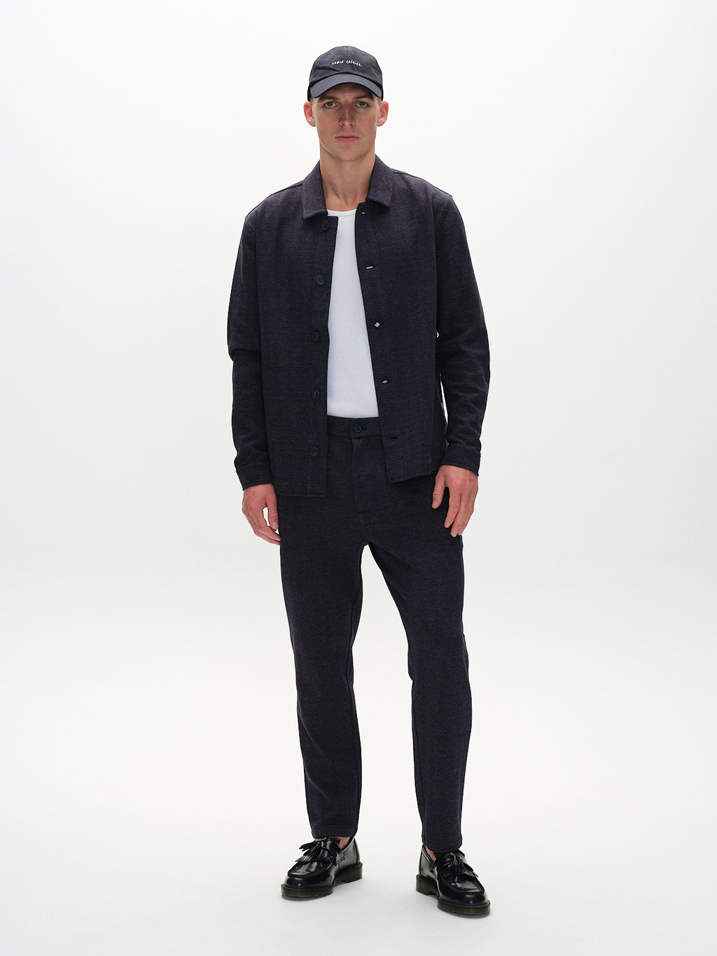 Against a white backdrop, a man sports a sophisticated navy look featuring Gabbas Monza Tweed pants. He pairs them with a button-up shirt, cap, black shoes, and layers it over a crisp white shirt for an added touch of refinement.